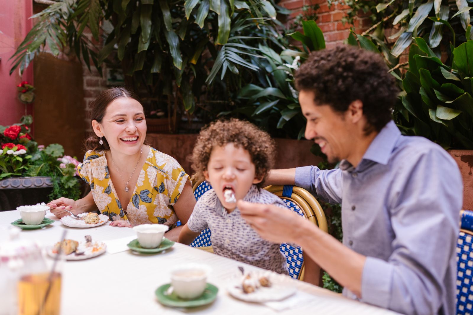 Parents laughing with their young son as he takes a bite at a restaurant patio table